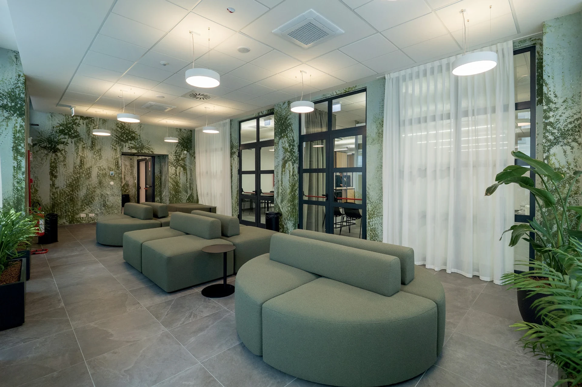 Area comune lounge con divani modulari verdi, piante tropicali e illuminazione soffusa. Palazzo Contarini student house, Udine. Arredamento su misura Tomasella Contract, 2025.
